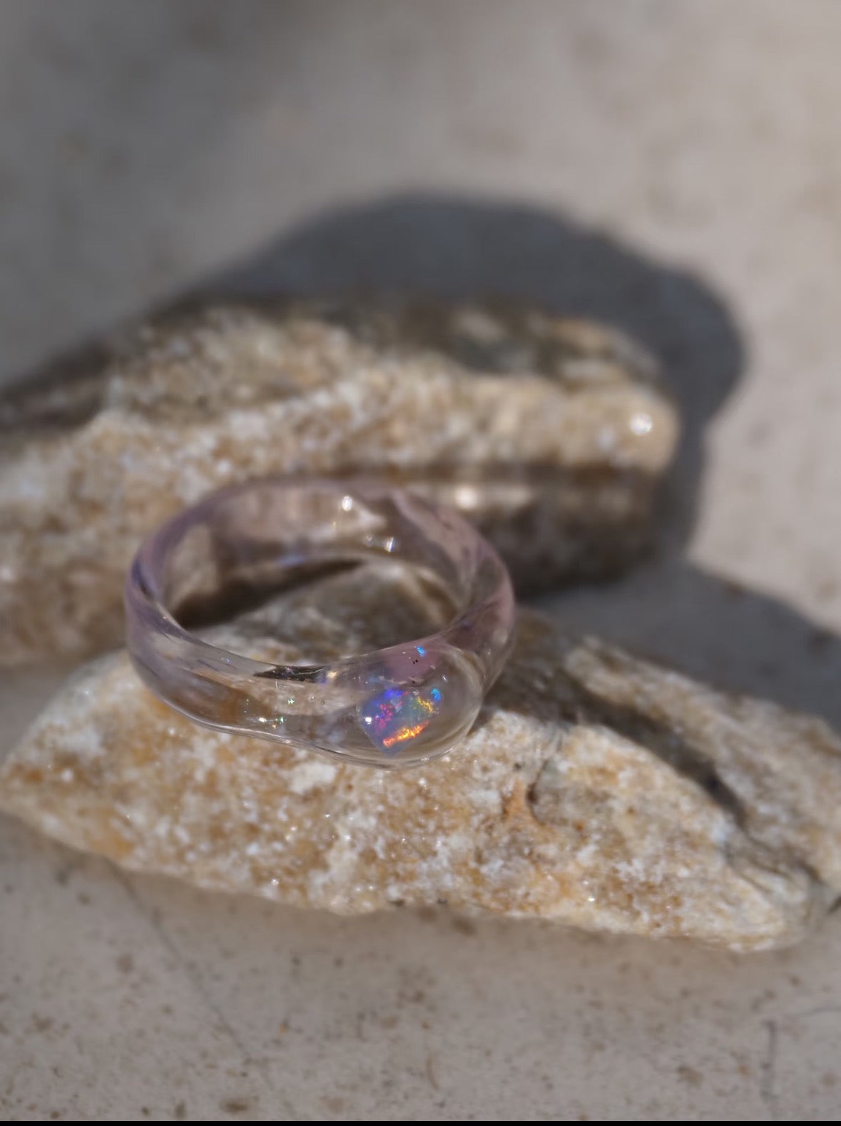 Clear hand-shaped glass ring with a lab-grown opal, resting on pale stone.